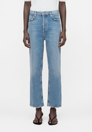 Femeie purtând blugi scurți, cu talie înaltă, albastru deschis, cu tiv nefinisat și sandale negre cu barete, stând pe un fundal alb.