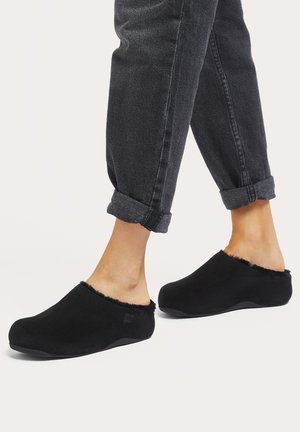 Black slip-on clogs with a soft fur lining at the collar and a rounded toe shape, set against gray cuffed denim pants.