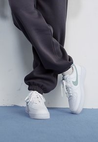 Bijele Nike Air Force 1 tenisice s tirkiznim akcentima, postavljene uz siva trenirka s elastičnim manžetama, na svijetloplavoj površini.