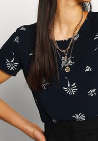 Haut bleu marine à motif floral blanc, manches courtes, texture côtelée. Colliers superposés en métal doré avec charms. Jupe noire à la taille.