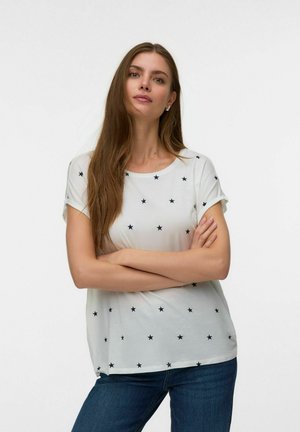Kvinde med langt brunt hår står med armene over kors iført en hvid T-shirt med sort stjernemønster og blå jeans foran ensfarvet baggrund.