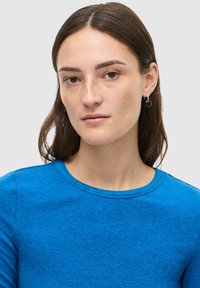 Blå, teksturert bomulls T-skjorte med rund hals. Modellen har på seg runde, svarte øredobber. Enkel design med nærbilde av øvre del.