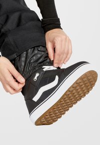 Zwarte hoge sneakers met witte accenten en een textuurrubber zool. Close-up van een hand die de sluiting bij de enkel aanpast.