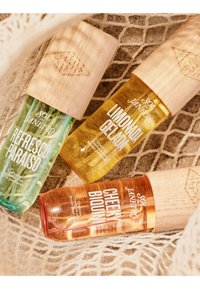 Tres mists de perfume Sol de Janeiro Cheirosa—Refresco Paraíso, Limonada Gelada y Cheeky Biquini—con tapones de madera sobre una tela beige texturizada.