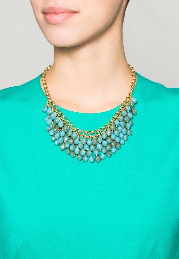 Femme portant un collier en chaîne dorée avec plusieurs perles turquoises pendantes sur un haut turquoise vif, cou et bas du visage visibles.