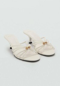 Sandales à talons blancs avec des lanières, dotées d'un petit talon, présentant une texture lisse et un accent doré central. Design simple et style ouvert à l'avant.