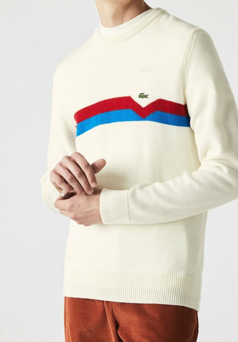 Personne portant un pull blanc avec des rayures en chevrons rouges et bleues et un petit logo crocodile vert sur la poitrine, associé à un pantalon marron.