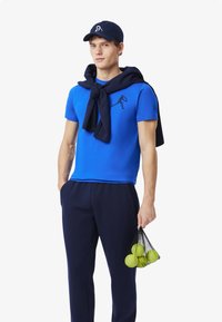 Sininen lyhythihainen paita, jossa on musta logo, tummansiniset housut ja samalla värillä varustettu lippis. Pidättelee verkko laukku, jossa on tennispalloja. Neule on heitetty hartioille.
