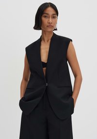 Schwarze ärmellose Blazer mit tiefem V-Ausschnitt, strukturierten Schultern und Vordertaschen. Hergestellt aus glattem Stoff mit einem minimalistischen Design.