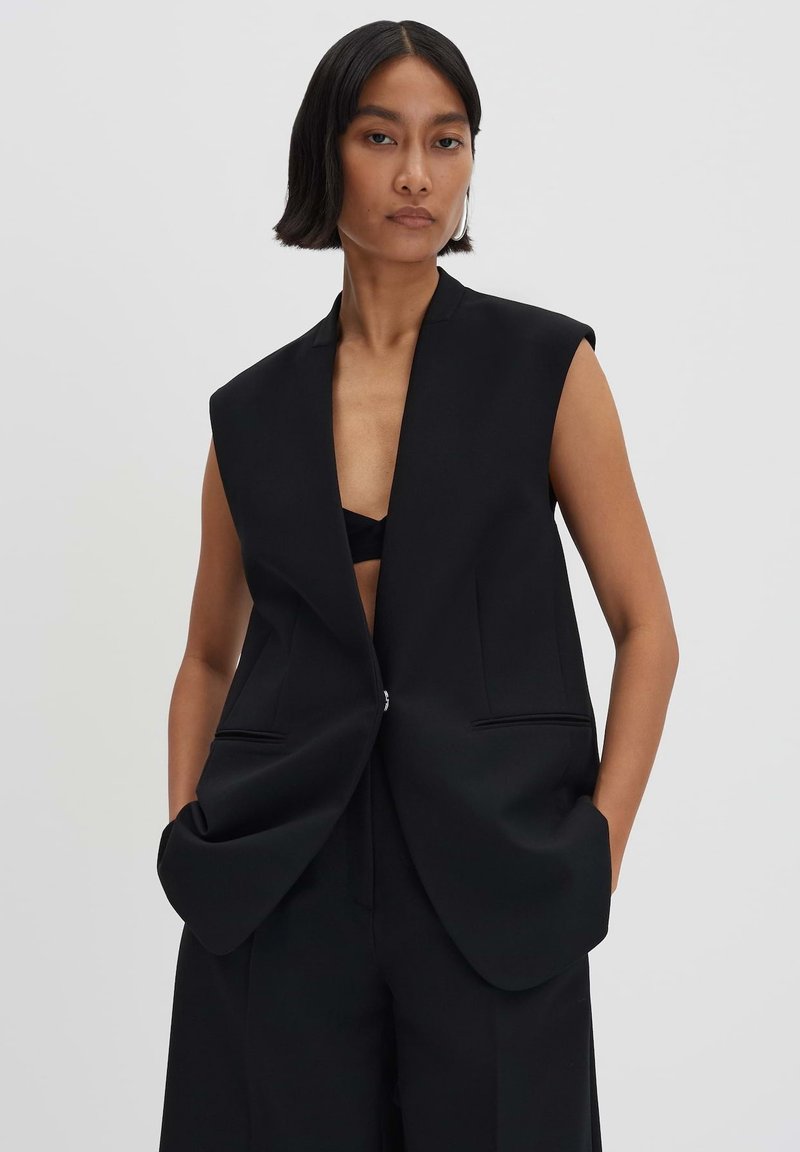 Schwarze ärmellose Blazer mit tiefem V-Ausschnitt, strukturierten Schultern und Vordertaschen. Hergestellt aus glattem Stoff mit einem minimalistischen Design.
