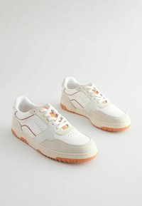 Białe i beżowe sneakersy z pomarańczowymi akcentami i teksturowanymi materiałami. Posiadają grubą podeszwę, perforowany czubek oraz kontrastujące panele boczne.