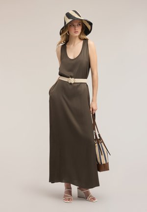 Vrouw draagt een mouwloze bruine maxi-jurk met een beige riem, gestreepte brede hoed, witte sandalen en houdt een gestreepte handtas vast.