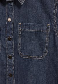 Mörkblå denimskjorta med en framficka, metallnitar på stolpen och kontrasterande gyllene sömmar. Slät textur.