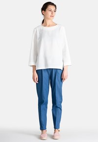 Krémbová blúzka s tromi štvrťovými rukávmi, skombinovaná s modrými džínsami. Model má na sebe svetloružové espadrilky. Jednoduchý, minimalistický dizajn.
