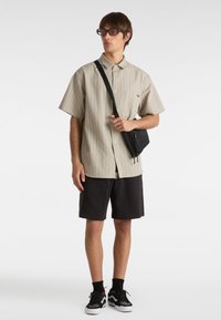 Camicia a maniche corte beige a strisce, pantaloni corti neri e borsa a tracolla nera. Indossa occhiali da sole rosa oversize e scarpe da ginnastica nere/grigie.