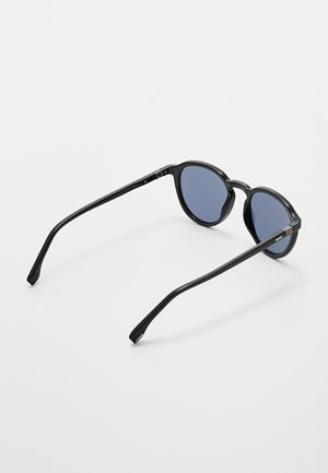 Occhiali da sole rotondi neri con lenti blu scuro che riposano su una superficie bianca, mostrati da dietro con un leggero angolo.