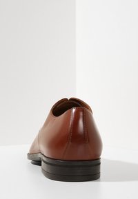 Chaussure de costume en cuir marron poli, vue de l'arrière, posée sur une surface blanche avec un fond minimaliste blanc.