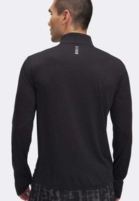 Man die met zijn rug naar de camera staat, gekleed in een zwart langemouwen T-shirt met een klein verticaal wit streep-logo op de bovenrug en een patroon op de broek.