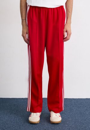 Homme portant un pantalon de survêtement rouge avec des bandes blanches sur les côtés et des baskets blanches, debout sur un tapis bleu.