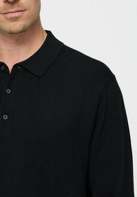 Mees, kes kannab musta pika varrukaga polo särki, on kujutatud kaelast kuni kesk torso, lihtsa heleda tausta ees.