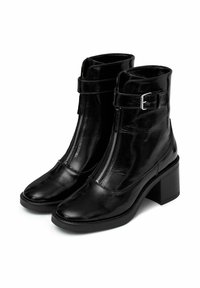 Botines de cuero negro hasta el tobillo con un alto tacón cuadrado, que cuentan con un cierre lateral de cremallera y una correa con detalle de hebilla. Textura brillante con punta redondeada.