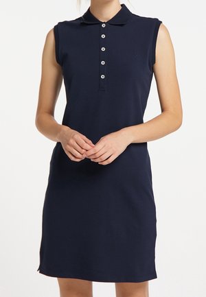 Robe polo sans manches bleu marine avec patte de boutonnage et petit logo ancre sur la poitrine, portée par une personne les mains jointes devant.