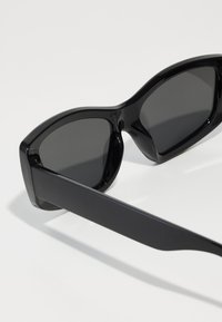 Black sunglasses with a glossy finish, featuring dark lenses and a thick, angular frame. The arm is streamlined and matte.
