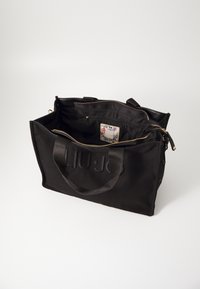Sac fourre-tout en tissu noir avec logo texturé, poignées en tissu dual, et fermeture éclair dorée, offrant un intérieur spacieux et doublé ainsi qu'une étiquette.