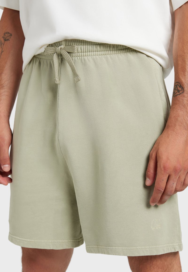 Short beige à cordon porté avec une chemise blanche, montrant les mains et les bras avec de petits tatouages à chaque poignet.
