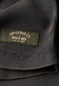 Ciemnoszary dzianinowy materiał z zieloną prostokątną metką, na której napisano "Superdry Co. Machined Dry Goods" i która jest do niej przyszyta.