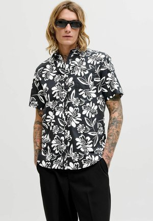 Homme tatoué portant une chemise à manches courtes noire et blanche à motifs floraux, un pantalon noir et des lunettes de soleil foncées, les mains dans les poches.