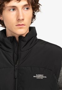 Svart pufferväst med hög krage och dragkedjestängning. Har vit logotext "ELEMENT SKATEBOARD CO. WORLD WIDE" på bröstet.