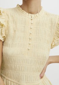 Hellgelbes Kleid mit einem gerafften Material, kurzärmligen Rüschen und fünf kleinen Knöpfen an der Vorderseite. Weicher, leichter Stoff.