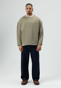 Olivengroen oversized sweatshirt met lange mouwen, gedragen over een wit t-shirt. Gecombineerd met losse donkere spijkerbroek en bruine laarzen.