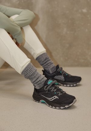 Osoba siedząca na podłodze, ubrana w jasnobeżowe spodnie, grube szare skarpety oraz czarne sportowe buty z akcentami w kolorze morskim i szarym.