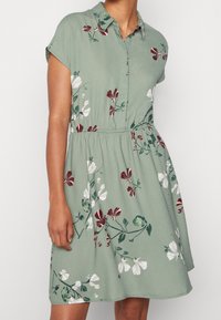 Kortärmad klänning med knappar i ljusgrönt tyg, med ett blommönster i rött, vitt och grönt, och resår i midjan.