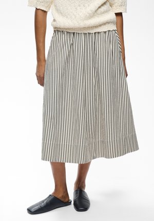 Persona che indossa un top in maglia color crema, una gonna midi a righe verticali bianche e nere e scarpe nere senza lacci, in piedi contro uno sfondo semplice.
