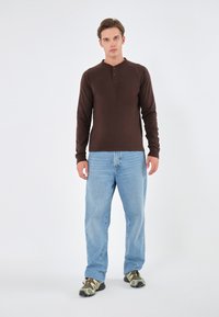Cămașă henley cu mânecă lungă de culoare maronie, cu guler butonat, combinată cu blugi lejeri de culoare albastru deschis și încălțări atletice cu model camuflat.