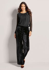 Zwarte geplooide broek van gestructureerde stof, gecombineerd met een vloeiende zwarte top met doorschijnende lange mouwen.
