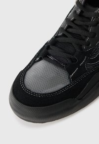 Black sneaker featuring a mix of black suede and gray mesh, with a textured rubber sole, black laces, and white stitching accents.
