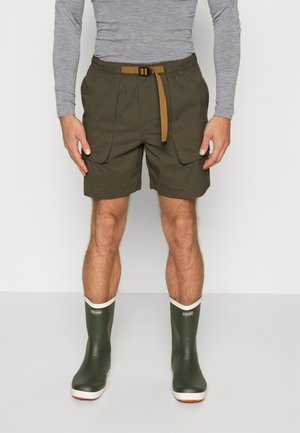 Homme portant un short cargo vert olive avec une ceinture beige, une chemise grise à manches longues et des bottes en caoutchouc vertes, debout sur un fond uni.