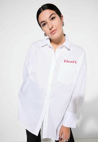 Camicia bianca oversize con bottoni, maniche lunghe e tasca a sinistra con scritta rossa che dice "Non farlo." Tessuto di cotone liscio.