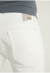Jeans de mezclilla blanca con un acabado texturizado. Presenta un bolsillo trasero con costuras diagonales y una etiqueta de marca de cuero beige etiquetada "CHASIN'".