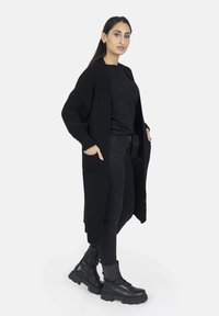 Schwarze Strickjacke mit langen Ärmeln und offener Front, kombiniert mit einem schwarzen Oberteil und engen Hosen, ergänzt durch schwarze Schnürstiefel.