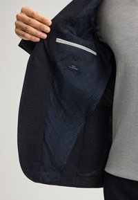 Marineblazer mit strukturierter Innenfutter. Ausgestattet mit einer grauen Tasche und einem Etikett mit der Aufschrift "Feinster italienischer Stoff." Glatte Oberfläche und maßgeschneiderte Passform.