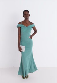 Rochie teal cu umeri descoperiți, cu dantelă la margine, corset ajustat și fustă fluidă. Modelul ține în mână o plic argintiu și poartă tocuri verzi.