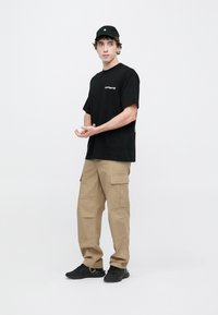 Czarny oversize'owy T-shirt z białym logo, beżowe spodnie typu cargo z bocznymi kieszeniami oraz czarne sneakersy, na jednolitym białym tle.