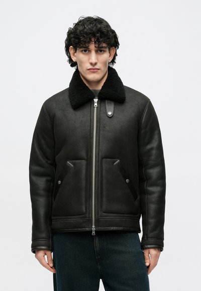 Juoda odinė striukė su minkštu juodu avies vilnos apykakle, užtrauktuku ir dviem įstrižomis priekinių kišenių. Lygi tekstūra, pritaikyta figūrai.