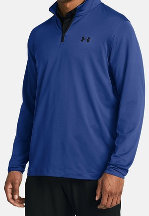 Homme portant un pull bleu à manches longues avec fermeture éclair quart de zip, un logo noir sur la poitrine et un pantalon noir, debout les bras détendus.