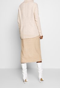 Beiger Strickpullover mit hohem Kragen, kombiniert mit einem karierten beige Midi-Rock. Das Outfit wird durch kniehohe weiße Stiefel mit Absatz vervollständigt.